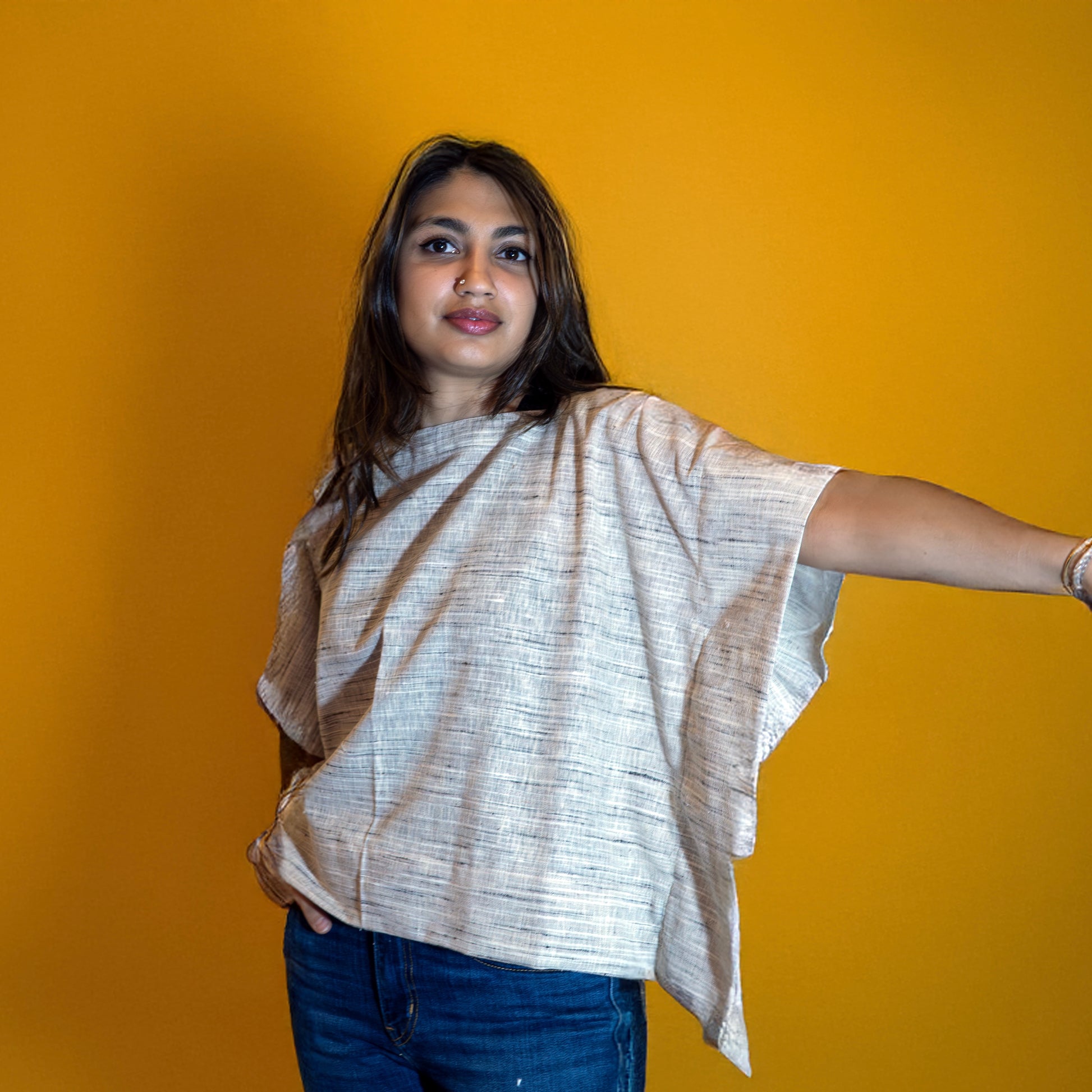 Woman wearing sustainable handwoven cotton poncho top by Surkh Amour in a relaxed fit, styled with denim, photographed against a yellow background.